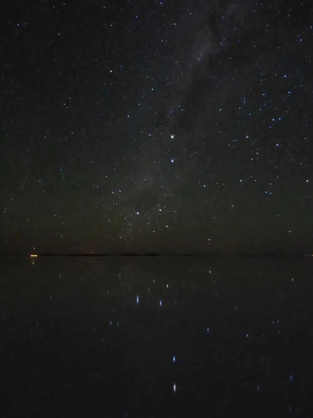 ウユニ塩湖の湖面に星空が映り込み、遠くに街の明かりが見える