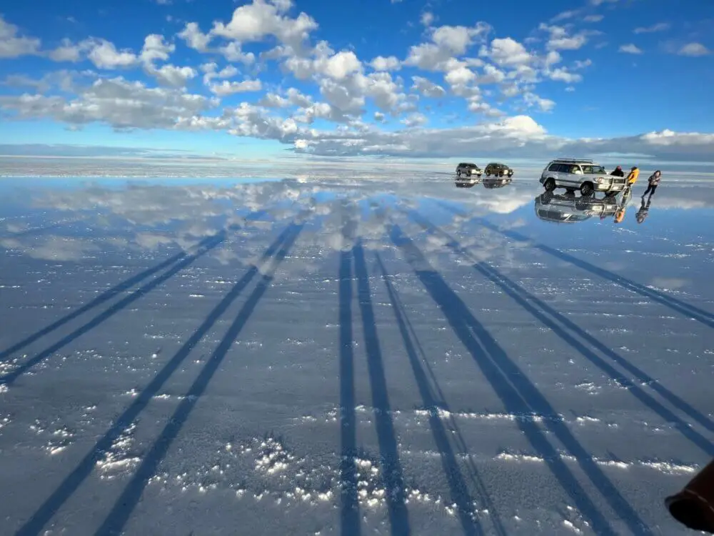 ウユニ塩湖の水面に空と雲が映り込み、車と人々の長い影が伸びる。