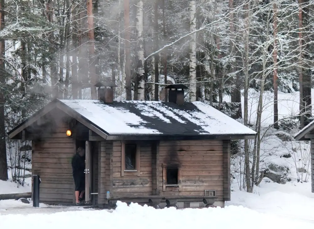 雪に囲まれた森の中、煙を上げる木造のサウナ小屋と人影