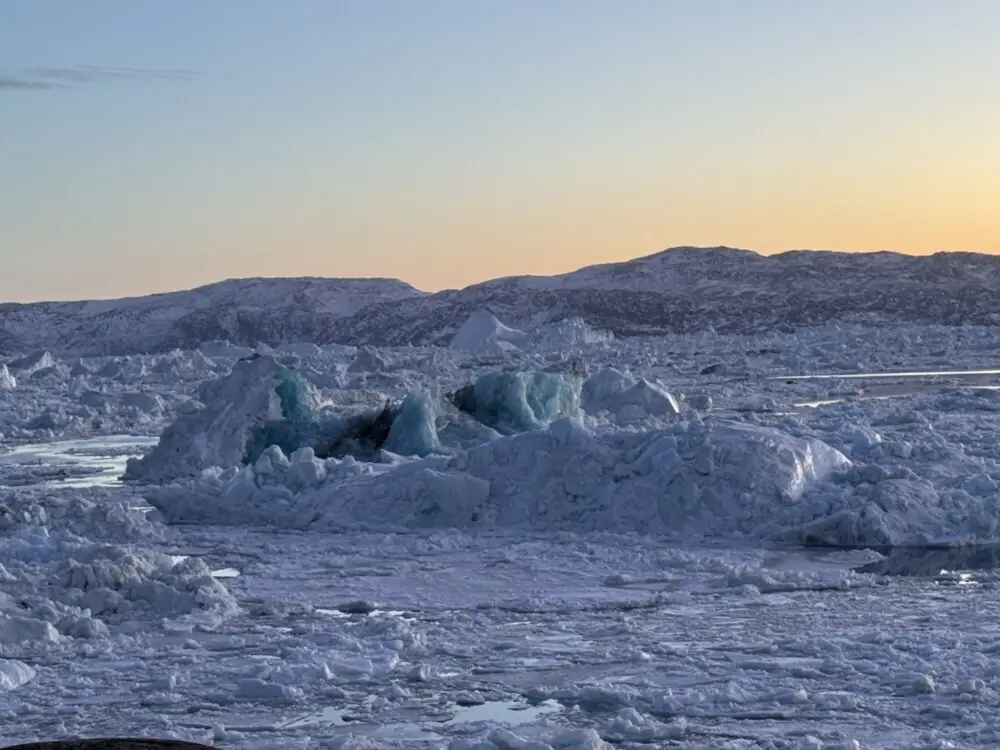 氷山が広がる水面に雪を抱く山々が連なる、夕暮れ時のグリーンランドの風景